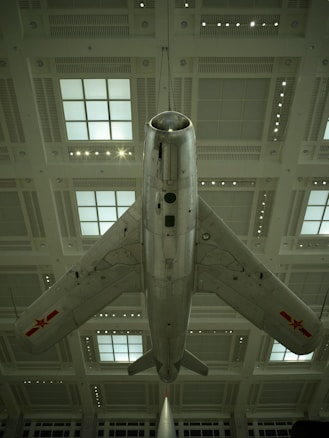 A vintage military jet airplane is suspended from the ceiling of a large indoor exhibition space. The aircraft has a sleek silver body with marked symbols on each wing, and it is viewed from below, emphasizing its aerodynamic shape and design features.