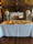 An elegant banquet setup is visible with round tables covered in white tablecloths, complemented by gold overlays. Tall vases filled with large, white floral arrangements are placed on a white counter, with a stone fireplace in the background. The setting gives an impression of a formal event venue with a rustic touch.
