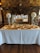 An elegant banquet setup is visible with round tables covered in white tablecloths, complemented by gold overlays. Tall vases filled with large, white floral arrangements are placed on a white counter, with a stone fireplace in the background. The setting gives an impression of a formal event venue with a rustic touch.
