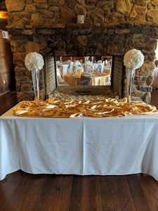 An elegant banquet setup is visible with round tables covered in white tablecloths, complemented by gold overlays. Tall vases filled with large, white floral arrangements are placed on a white counter, with a stone fireplace in the background. The setting gives an impression of a formal event venue with a rustic touch.
