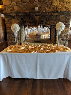 An elegant banquet setup is visible with round tables covered in white tablecloths, complemented by gold overlays. Tall vases filled with large, white floral arrangements are placed on a white counter, with a stone fireplace in the background. The setting gives an impression of a formal event venue with a rustic touch.