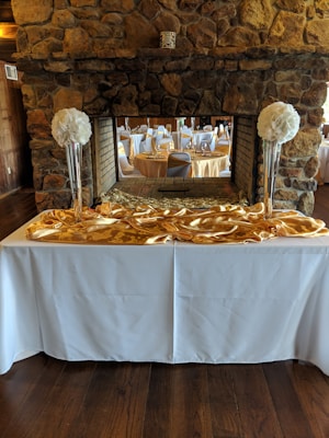An elegant banquet setup is visible with round tables covered in white tablecloths, complemented by gold overlays. Tall vases filled with large, white floral arrangements are placed on a white counter, with a stone fireplace in the background. The setting gives an impression of a formal event venue with a rustic touch.