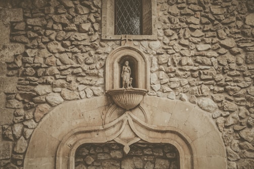 An ornate stone wall features a carved niche housing a small religious statue above a decorative arch. The textured stones create a rustic and ancient appearance, with a small window featuring diamond-shaped panes above the niche.