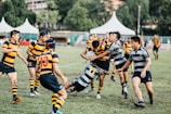 A group of young men are playing rugby on a grassy field. Two players in blue and white striped uniforms are tackling a player in a black and yellow striped uniform, who is holding the rugby ball. Other players, also wearing black and yellow uniforms, are nearby watching the play. In the background, there are white tents, trees, and a partially visible building.