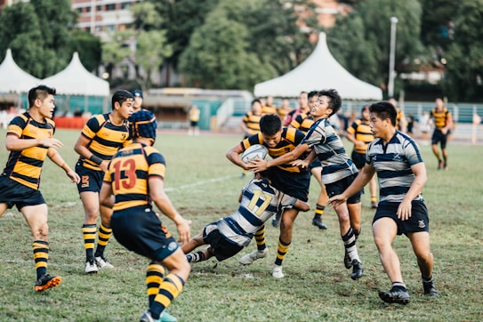 A group of young men are playing rugby on a grassy field. Two players in blue and white striped uniforms are tackling a player in a black and yellow striped uniform, who is holding the rugby ball. Other players, also wearing black and yellow uniforms, are nearby watching the play. In the background, there are white tents, trees, and a partially visible building.