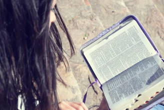A person with long dark hair focuses on reading a book with detailed text. The book has a colorful cover, and the scene is set outdoors on a sandy or stony surface.