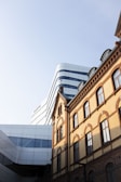 A juxtaposition of an old and new building. The old building has a classic architectural style with arched windows and an ornate facade. The new building has a sleek, modern design with curved lines and reflective surfaces. The sky is clear and provides a clean backdrop for the buildings.