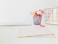 A minimalist desk with a laptop, a pink coffee cup, and a small vase of dried flowers, all in soft neutral shades.