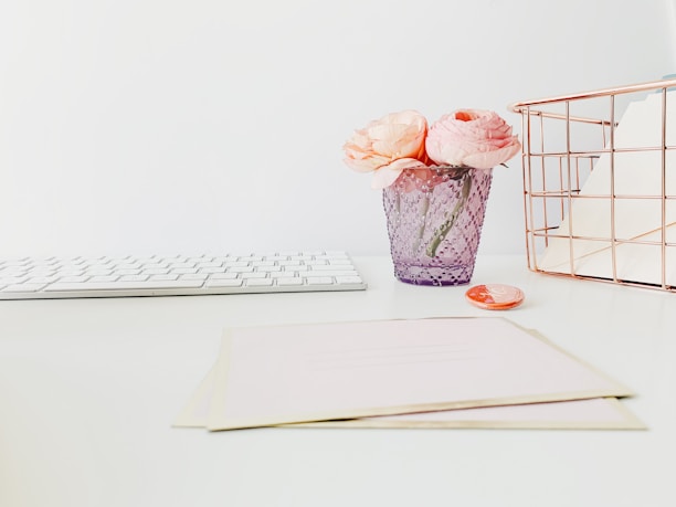 A minimalist workspace with a white keyboard on the left, a lilac vase containing light pink flowers in the center, and a rose gold wire desk organizer on the right. A blush pink envelope and a small round object are placed nearby on the desk, all set against a white background.