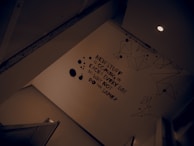 A wall is adorned with a motivational statement in bold black text. There are geometric line drawings and black ink splatter designs around the text. The scene is lit by a ceiling light, creating a modern and somewhat introspective atmosphere.