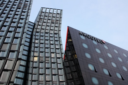 Modern architecture featuring two tall skyscrapers with glass facades reflecting the sky. The building on the right has a distinctive red trim and a sign labeled 'ARCOTEL'. The sky is clear with a light blue hue.