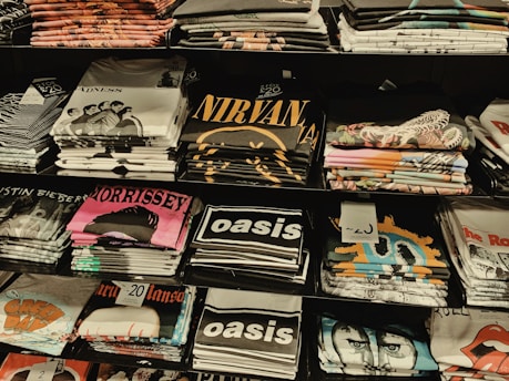 Several stacks of folded band t-shirts are displayed on shelves, featuring various music groups and artists such as Madness, Nirvana, Morrissey, Oasis, and others. Each stack is neatly arranged and some shirts have visible price tags.