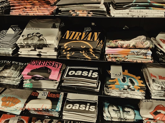 Several stacks of folded band t-shirts are displayed on shelves, featuring various music groups and artists such as Madness, Nirvana, Morrissey, Oasis, and others. Each stack is neatly arranged and some shirts have visible price tags.
