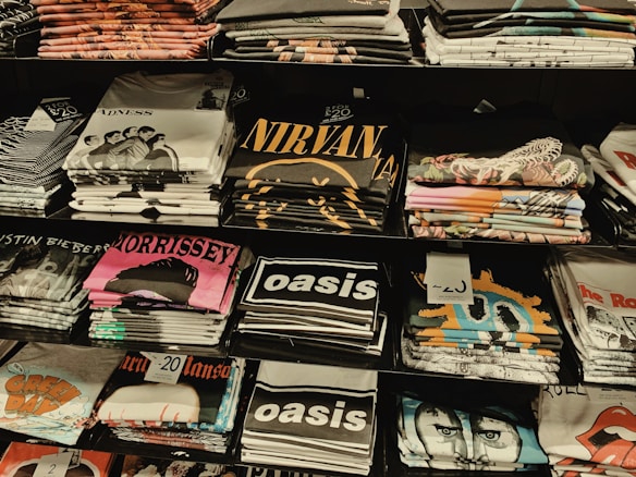 Several stacks of folded band t-shirts are displayed on shelves, featuring various music groups and artists such as Madness, Nirvana, Morrissey, Oasis, and others. Each stack is neatly arranged and some shirts have visible price tags.