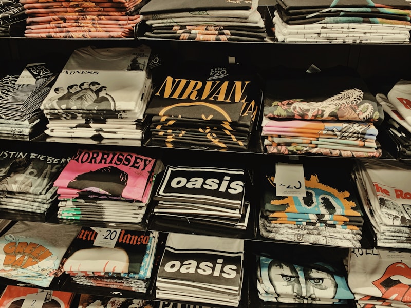 Several stacks of folded band t-shirts are displayed on shelves, featuring various music groups and artists such as Madness, Nirvana, Morrissey, Oasis, and others. Each stack is neatly arranged and some shirts have visible price tags.