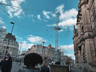 A scenic urban setting featuring historical architecture and modern elements. Tall buildings with intricate designs line the street, while a tall amusement park ride elevates above the scene with people seated in swing chairs. The sky is a bright blue with scattered clouds, and pedestrians are strolling along the pathways.