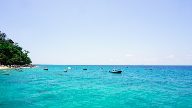 A serene coastal scene features crystal-clear turquoise waters dotted with small boats. On the left side, a lush green hillside with rocky edges gently touches the coastline.