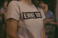A person wearing a white t-shirt with black lettering, standing among a group of people. The text on the shirt reads 'EMME VIBI'. The background is blurred, with hints of people and possibly other objects. A small heart tattoo is visible on their arm.