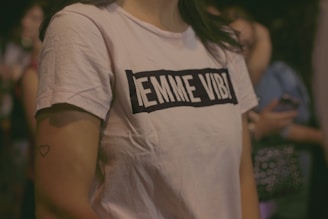 A person wearing a white t-shirt with black lettering, standing among a group of people. The text on the shirt reads 'EMME VIBI'. The background is blurred, with hints of people and possibly other objects. A small heart tattoo is visible on their arm.