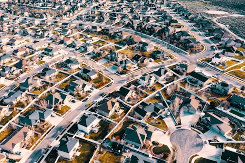 An aerial view of a suburban neighborhood displaying a network of streets and houses with neatly organized lawns and backyards. The landscape is characterized by a sprawling layout with similar-looking houses throughout the community.