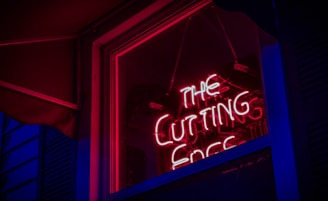 A brightly lit neon sign with the words 'THE CUTTING EDGE' is displayed inside a window, creating a striking contrast against the dark surroundings. The neon light casts a red and pink glow, which reflects on the glass pane and the structures nearby.