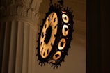 An ornate, circular clock with illuminated figures and designs is mounted on a fluted, classical-style pillar. The clock features silhouettes of various creatures and mythical beings around its edges, set against a warm, glowing background.