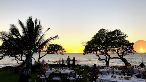 Tables are set up on a grassy area by the ocean at sunset, surrounded by palm trees and illuminated by a large glowing sphere. People are gathered at the tables, creating a festive atmosphere.