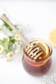 A jar filled with honey and a wooden honey dipper rests on top, adorned with small white flowers. Inside the jar, slices of lemon are visible, adding a fresh element. Surrounding the jar are delicate, small white flowers and green foliage, giving a natural and calming vibe.
