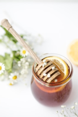 A jar filled with honey and a wooden honey dipper rests on top, adorned with small white flowers. Inside the jar, slices of lemon are visible, adding a fresh element. Surrounding the jar are delicate, small white flowers and green foliage, giving a natural and calming vibe.