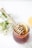 A jar filled with honey and a wooden honey dipper rests on top, adorned with small white flowers. Inside the jar, slices of lemon are visible, adding a fresh element. Surrounding the jar are delicate, small white flowers and green foliage, giving a natural and calming vibe.