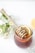 A jar filled with honey and a wooden honey dipper rests on top, adorned with small white flowers. Inside the jar, slices of lemon are visible, adding a fresh element. Surrounding the jar are delicate, small white flowers and green foliage, giving a natural and calming vibe.
