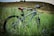 A mountain bike is parked in tall grass in front of a gentle hill. The bike has a sleek black and gray frame with some red and yellow accents. There is a small device mounted on the handlebars and the scene appears to be in an outdoor, natural setting.