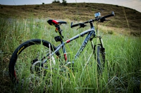 A mountain bike is parked in tall grass in front of a gentle hill. The bike has a sleek black and gray frame with some red and yellow accents. There is a small device mounted on the handlebars and the scene appears to be in an outdoor, natural setting.