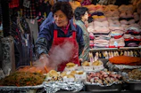 A woman wearing a red apron is cooking food at a busy market stall. Various types of food, such as noodles, skewers, and pastries, are laid out on trays, some covered in foil. In the background, stacks of clothes and folded garments are displayed, with another woman in a patterned jacket partially in view.