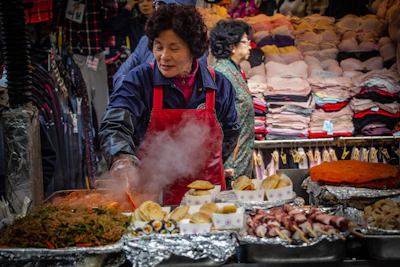 A woman wearing a red apron is cooking food at a busy market stall. Various types of food, such as noodles, skewers, and pastries, are laid out on trays, some covered in foil. In the background, stacks of clothes and folded garments are displayed, with another woman in a patterned jacket partially in view.