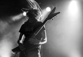 A musician is energetically playing an electric guitar on stage, with long hair flowing wildly. The scene is dramatic with stage lights creating a high-contrast effect, emphasizing the intensity of the performance.