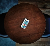 A smartphone displaying a colorful app interface lies at the center of a round wooden table. The dark wood grain contrasts with the bright phone screen. The table is surrounded by a couple of dark chairs positioned on a wooden floor.