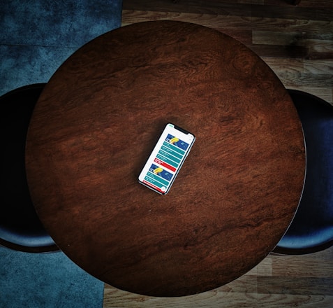 A smartphone displaying a colorful app interface lies at the center of a round wooden table. The dark wood grain contrasts with the bright phone screen. The table is surrounded by a couple of dark chairs positioned on a wooden floor.