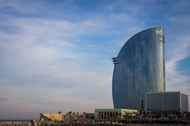 A tall, modern building with curved architecture stands prominently against the backdrop of a partly cloudy sky. The building has a sleek, reflective glass exterior and features a large 'W' logo at the top. At the base of the building, there are several palm trees and smaller structures.