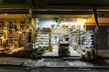 A well-lit store with a large variety of products displayed on shelves both inside and outside. The store's signage reads 'Amalthia's' and offers a range of food, drinks, beauty products, and other items. There are hanging lamps and potted plants near the entrance, contributing to a welcoming atmosphere.