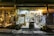 A well-lit store with a large variety of products displayed on shelves both inside and outside. The store's signage reads 'Amalthia's' and offers a range of food, drinks, beauty products, and other items. There are hanging lamps and potted plants near the entrance, contributing to a welcoming atmosphere.