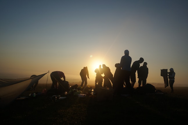 A group of people silhouetted against the rising or setting sun, engaged in various activities near what seems to be a camping setup. The sky is a gradient of warm and cool tones, and the horizon is softly visible in the distance.