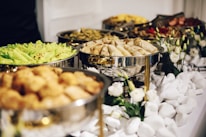 A buffet table with a variety of food items displayed in silver chafing dishes. Fresh greens, spring rolls, and other dishes are garnished with white flowers and surrounded by white decorative stones. The setup is elegant and organized, likely prepared for an event.