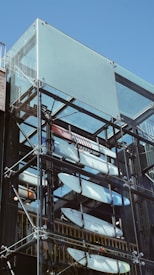 A modern, transparent structure displaying multiple surfboards inside, with metal supports and glass panels reflecting the clear blue sky.