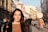 A young woman with long dark hair stands prominently in the foreground on a busy city street. She is wearing a brown top and a dark coat. The background is filled with people walking and engaging in various activities. The buildings are old and ornate, suggesting a historical or culturally rich area. A sign in Russian script is visible, indicating the location may be in a Russian-speaking country. The light suggests it's late afternoon or early evening with a warm, golden glow.