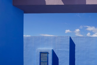 A vibrant blue wall with a small window and shutter casts a shadow against a clear sky. Clean lines and strong geometric shapes create a minimalist aesthetic.