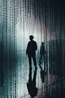 A silhouetted figure walks through a space filled with hanging, illuminated strands that create a shimmering, ethereal effect. The background is filled with vertical lines of light, and the reflective surface on the floor mirrors the figure and the lights, enhancing the surreal atmosphere.