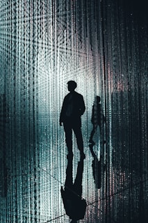 A silhouetted figure walks through a space filled with hanging, illuminated strands that create a shimmering, ethereal effect. The background is filled with vertical lines of light, and the reflective surface on the floor mirrors the figure and the lights, enhancing the surreal atmosphere.