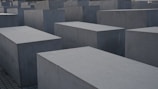 Large, rectangular concrete blocks arranged in a grid pattern, forming an abstract architectural landscape. The blocks are evenly placed on a paved ground, creating a sense of uniformity and structure.