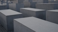 Large, rectangular concrete blocks arranged in a grid pattern, forming an abstract architectural landscape. The blocks are evenly placed on a paved ground, creating a sense of uniformity and structure.